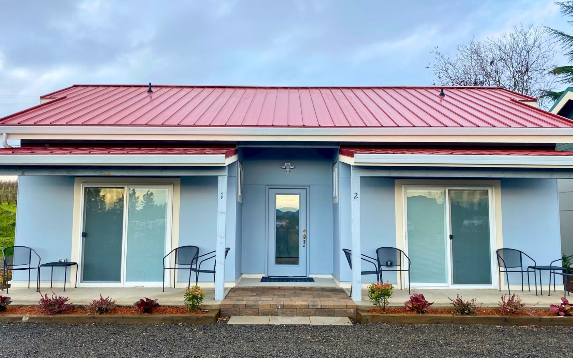 The image shows a small blue building with a red roof, two sliding glass doors, chairs on the porch, and plants in front.