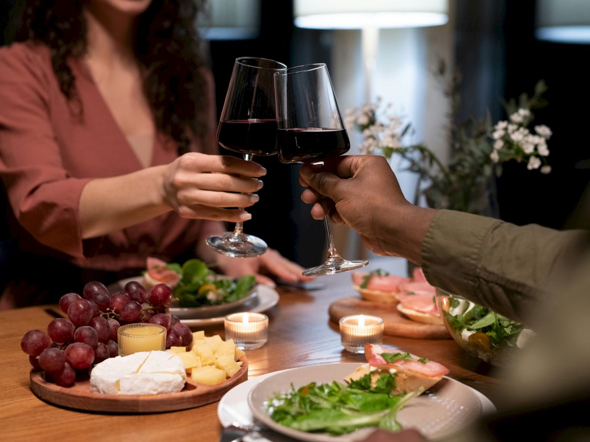Two people clink wine glasses over a table with cheese, grapes, and salad in a warm, candlelit setting.