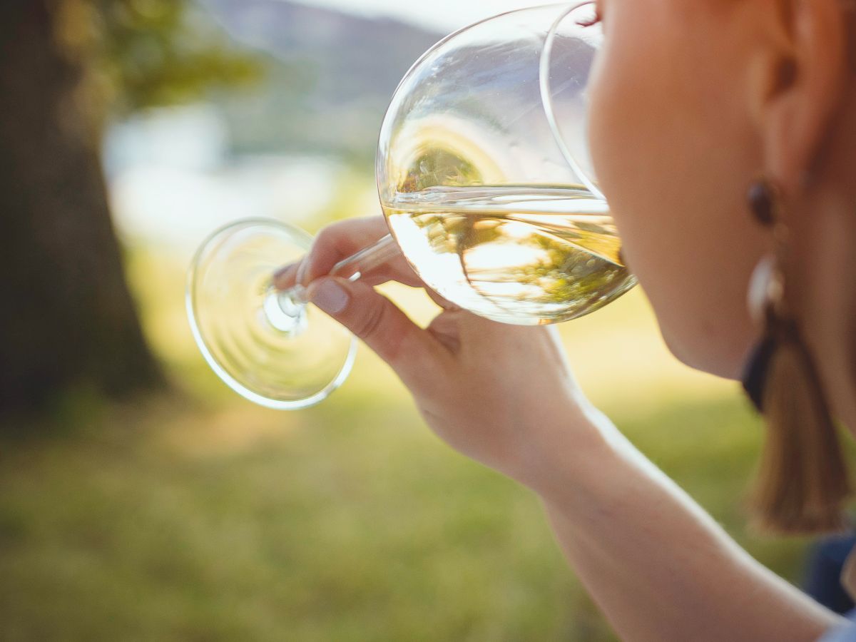 A person enjoys a glass of white wine outdoors, sitting on grass with trees and a blurry background, captured in warm daylight.