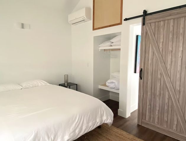 A bedroom with a bed, wooden sliding door, and open closet with shelves and hangers. Minimalist design with white walls and wooden flooring.