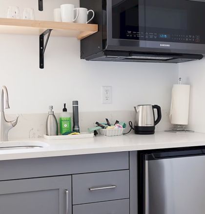 Small kitchen with a sink, gray cabinets, microwave, mini-fridge, kettle, and shelf with mugs. Paper towel roll and soap are visible.