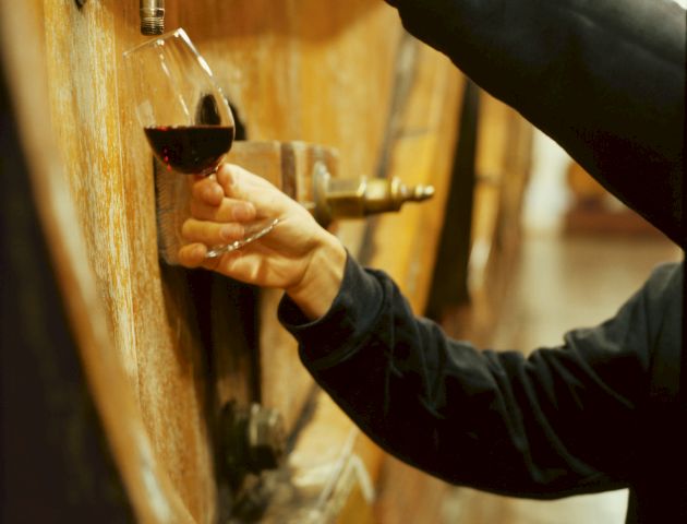 A person is pouring red wine from a large wooden barrel into a glass, using a spout.