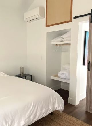 A bedroom with a bed, wooden sliding door, and open closet with shelves and hangers. Minimalist design with white walls and wooden flooring.