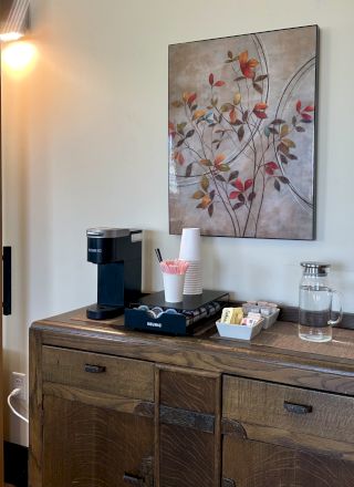 A wooden sideboard with a coffee maker, cups, water bottle, and condiments sits below floral artwork next to a sliding door.