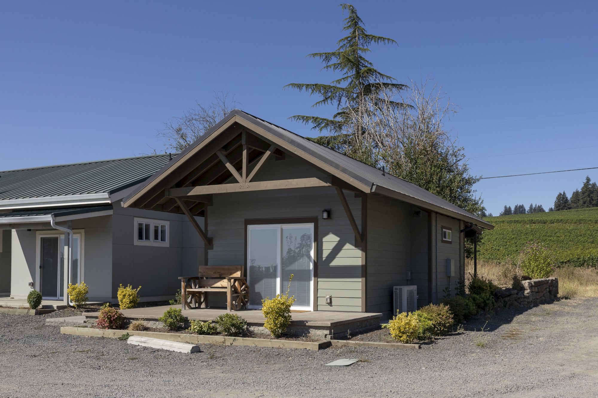A modern house exterior with dark metal roof, porch beams, windows, shrubs, and a tall evergreen tree against a clear blue sky.