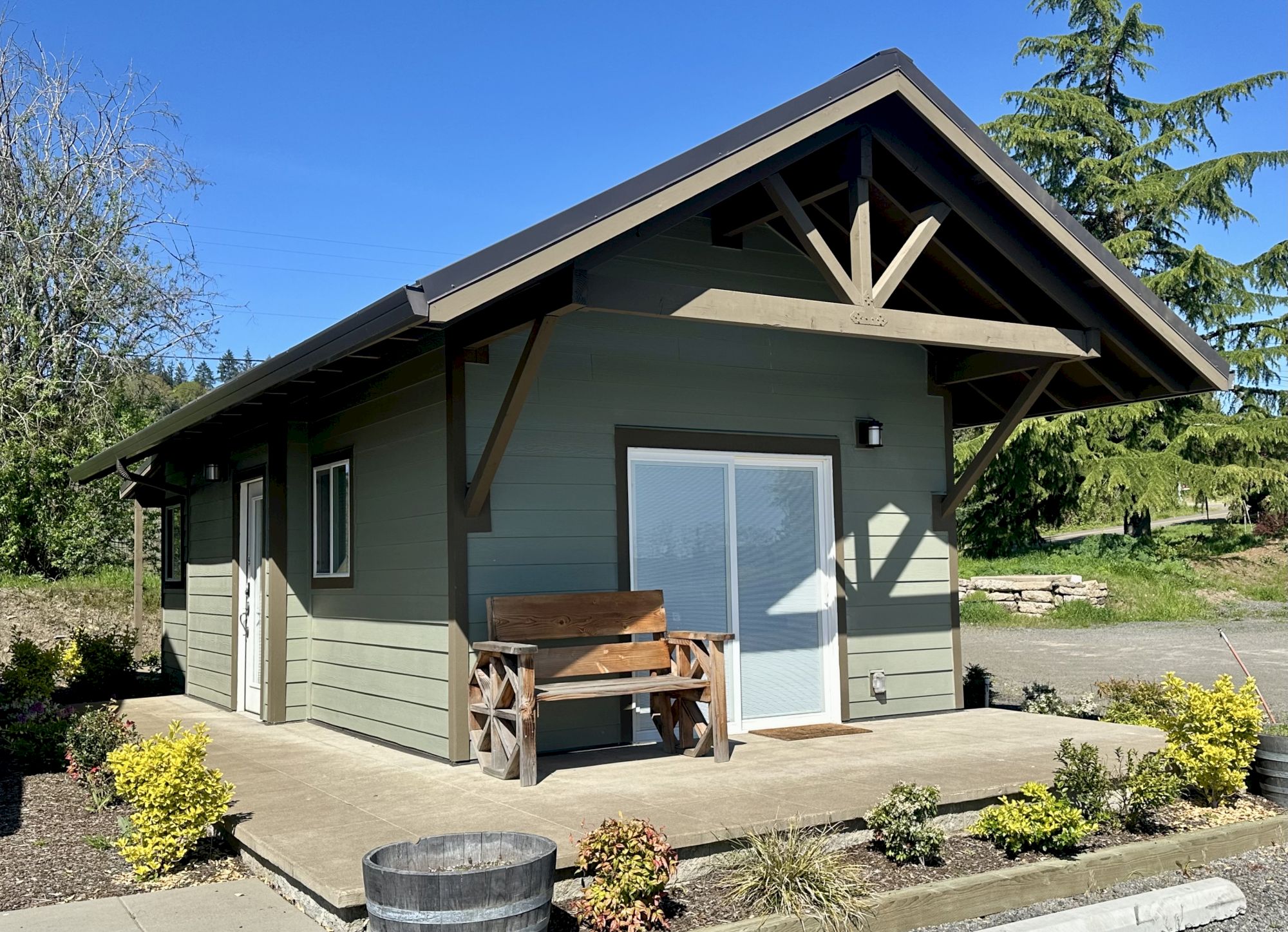 A small green cabin with a sloped roof, a wooden bench on the porch, sliding doors, and a tidy garden in front, under a bright blue sky.