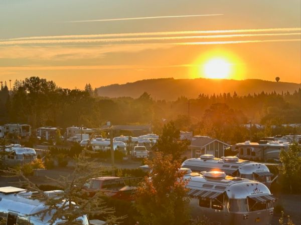 Sunset over an RV/camper park with many motorhomes, trees, and a misty hillside glowing in the distance.