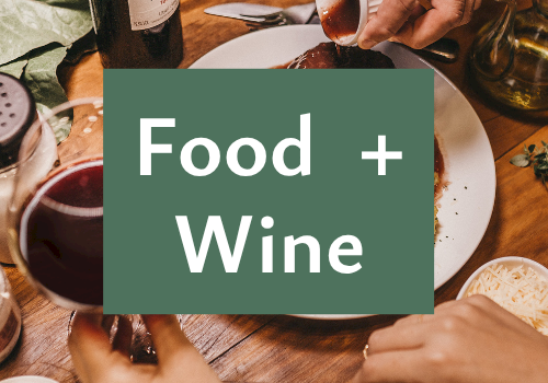 A hand pours red wine at a shared dinner table with food, cheese, and glasses.