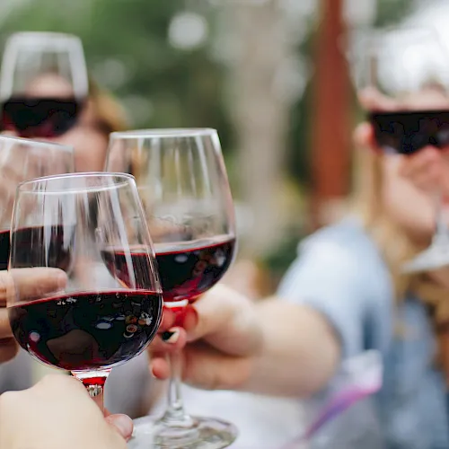 Friends clink glasses of red wine on a patio, smiling in the blurred background.