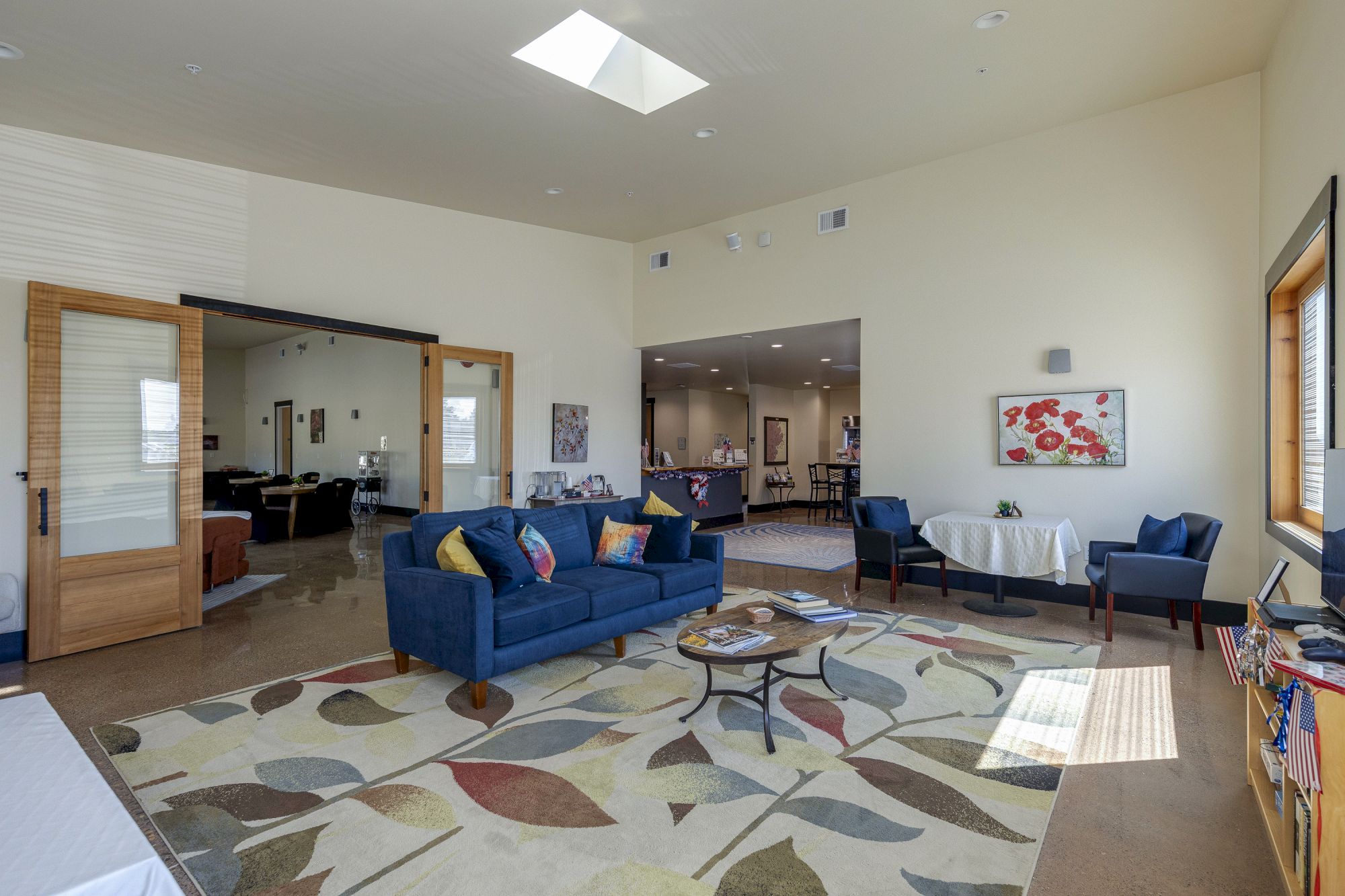 A bright living room with a blue sectional, patterned rug, coffee table, wall art, and an open view into the kitchen.