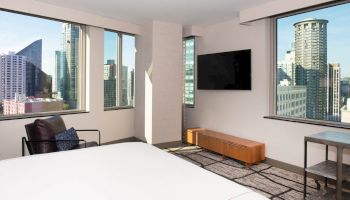 A modern hotel room with large windows, a bed, chair, TV, and views of urban skyscrapers and buildings outside.