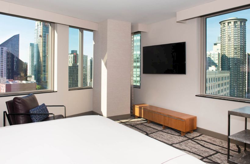 A modern hotel room with large windows, a bed, chair, TV, and views of urban skyscrapers and buildings outside.