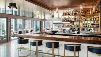 A modern bar with a wooden ceiling, stocked shelves, and stylish seating, set against large windows offering a city view.