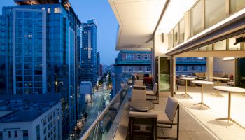 A modern rooftop terrace with tables and chairs overlooks a city street lined with tall buildings during twilight, creating a serene atmosphere.