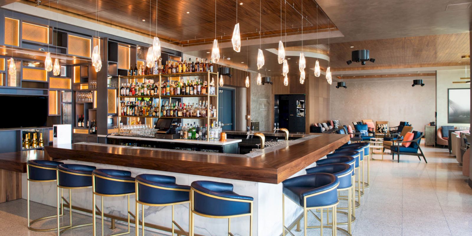 A modern bar with a wooden counter, blue chairs, hanging lights, and shelves of liquor bottles, set in a spacious, stylish interior.
