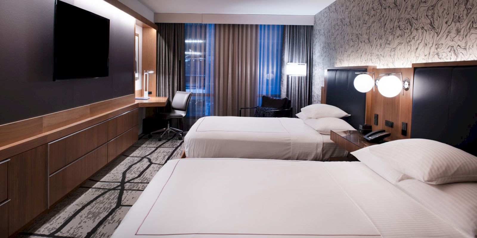 A modern hotel room with two beds, a desk with a chair, a wall-mounted TV, and patterned carpet and wallpaper.