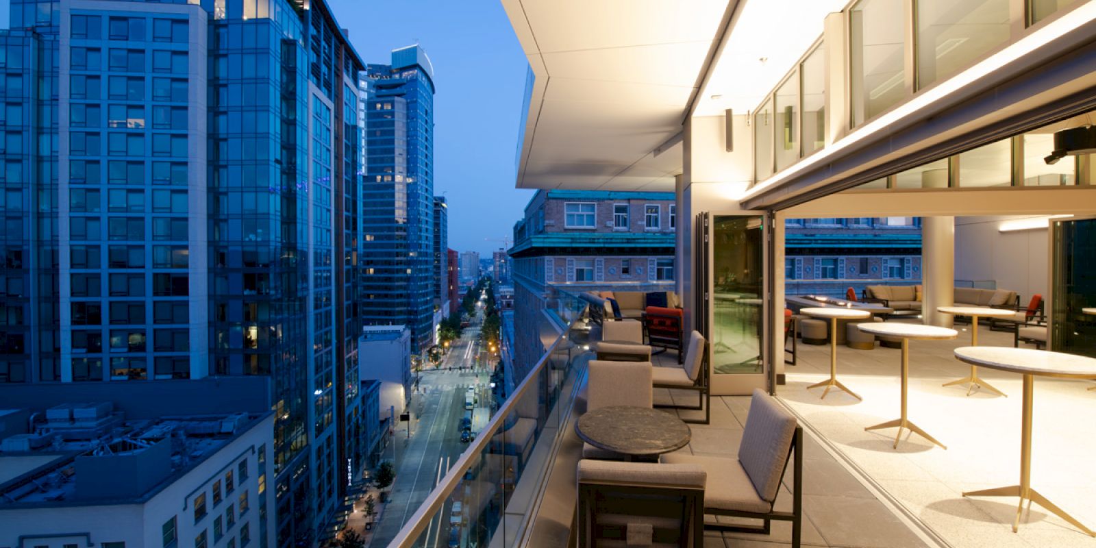 A modern cityscape at dusk with a sleek balcony view, featuring outdoor seating and nearby tall buildings along a street.