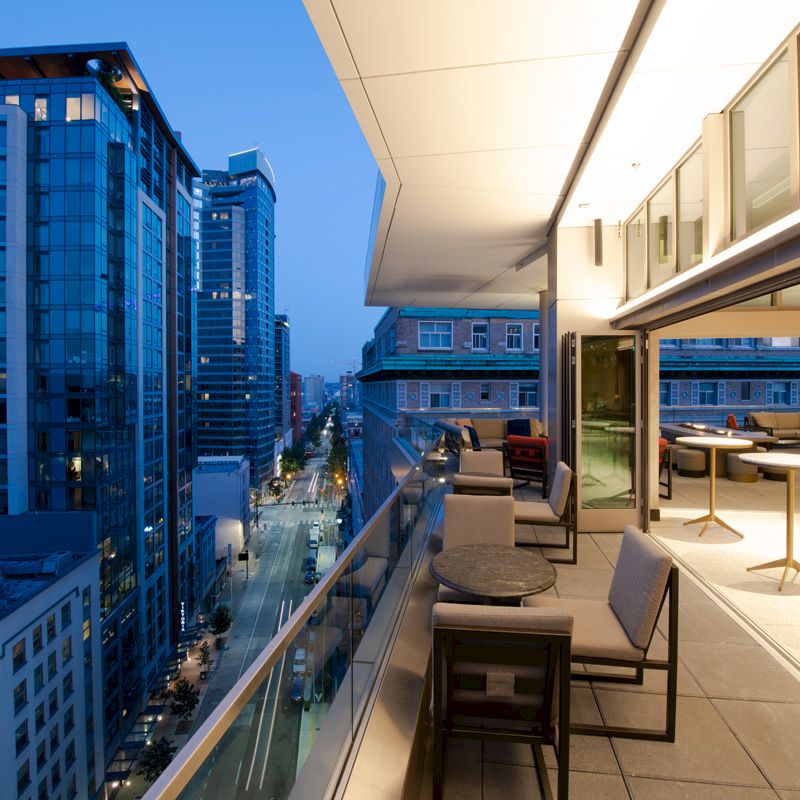The image shows a modern cityscape with tall buildings and a stylish balcony featuring tables and chairs, overlooking a street below.