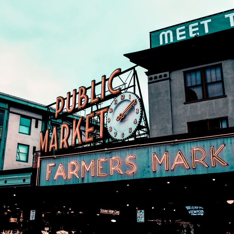 The image shows the entrance of a famous public market with neon signs reading "Public Market" and "Farmers Market" under a cloudy sky.