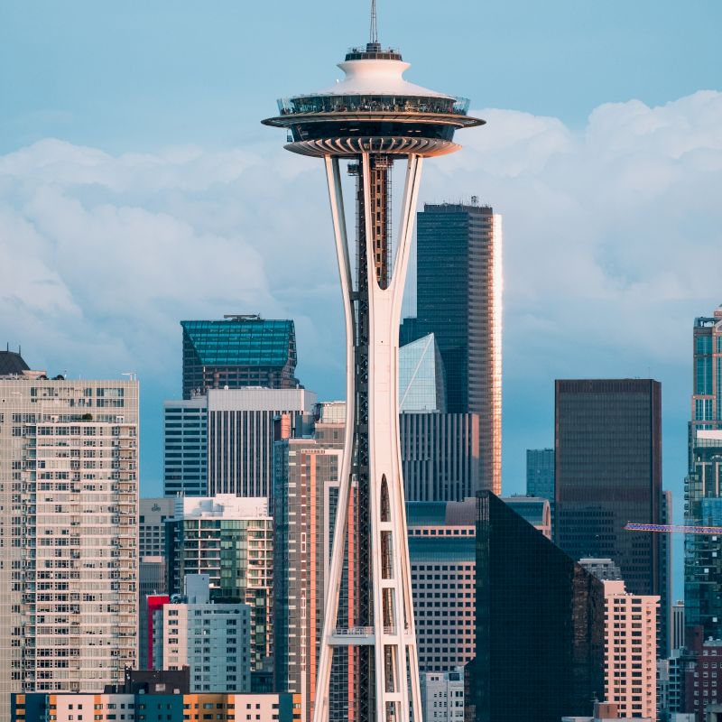 The image shows a city skyline with a white tower featuring a saucer-shaped top, surrounded by various tall buildings under a blue sky.