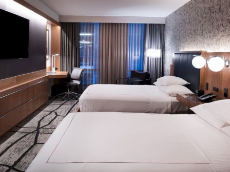 The image shows a modern hotel room with two beds, a TV, a work desk, and a large window with curtains.
