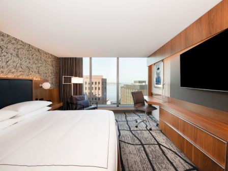 A modern hotel room featuring a large bed, wall-mounted TV, desk with a chair, and a window with cityscape views.