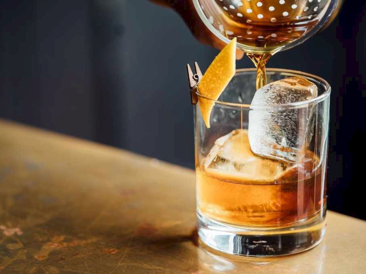 A cocktail is being poured into a glass with ice and a lemon twist garnish on a wooden surface.