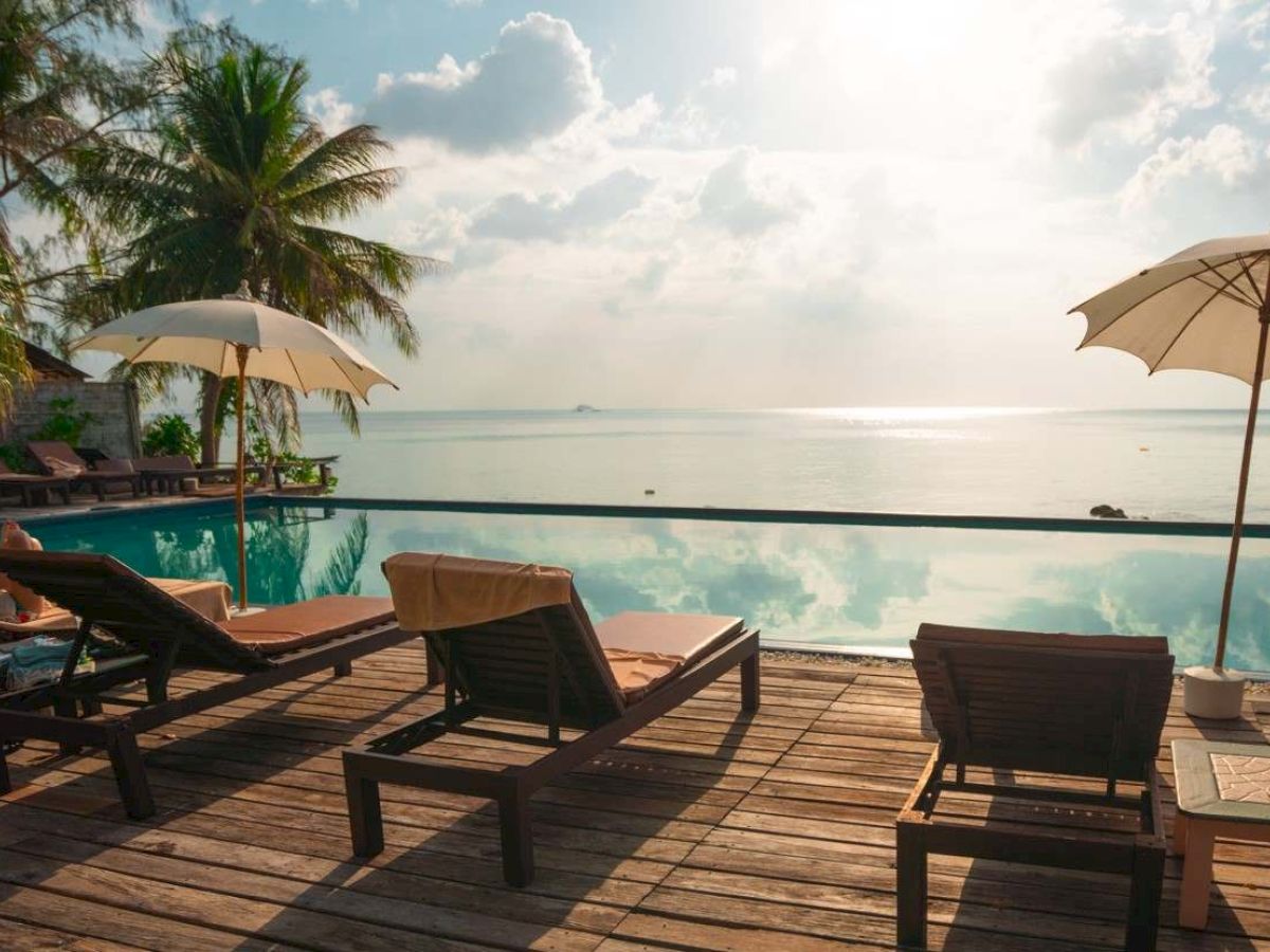 The image shows a tropical beach scene with lounge chairs, umbrellas, palm trees, and a calm ocean view under a partly cloudy sky.