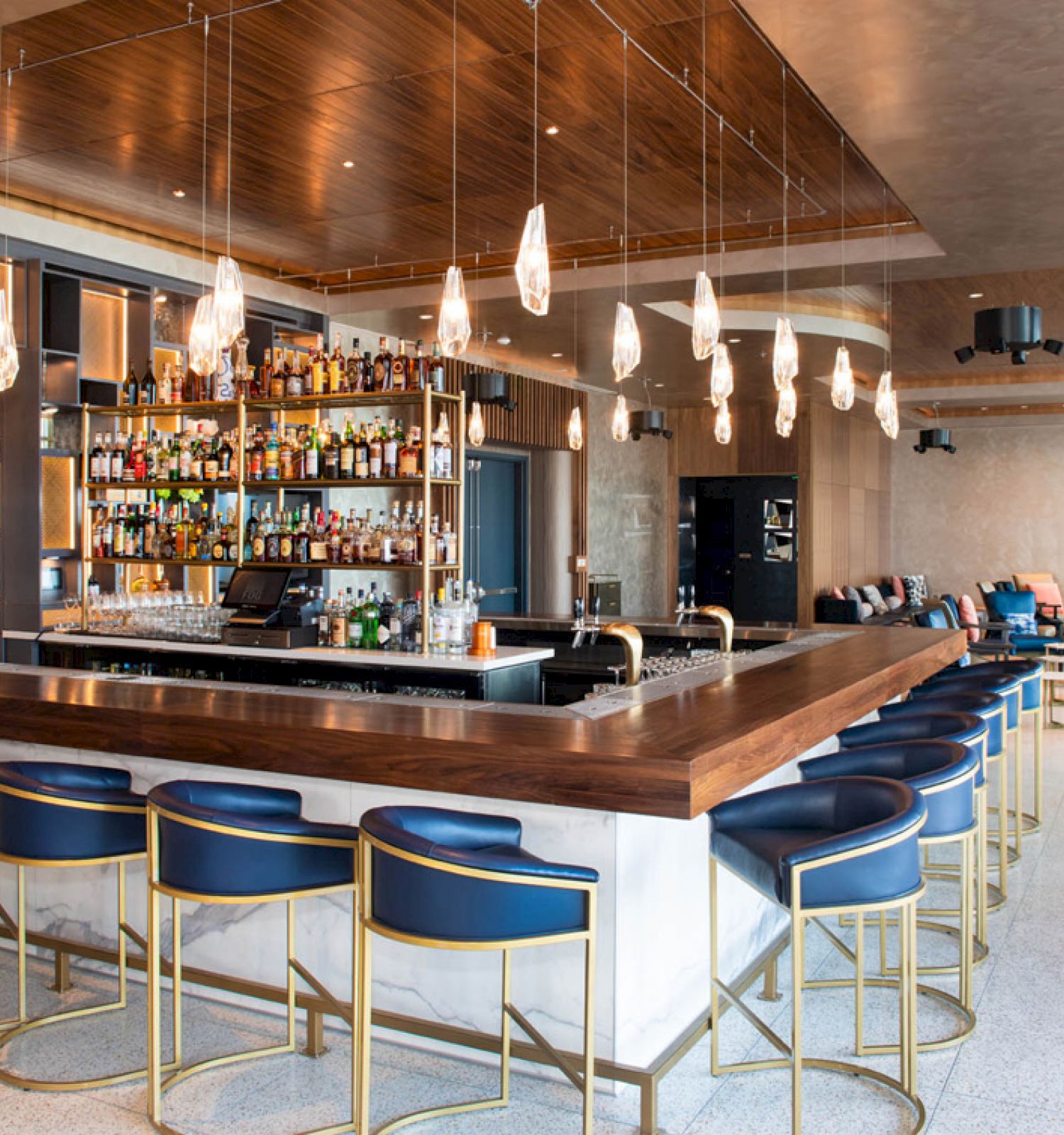 A modern bar with a wooden counter, blue stools, stylish lighting, and a well-stocked back bar in an open, chic lounge setting.