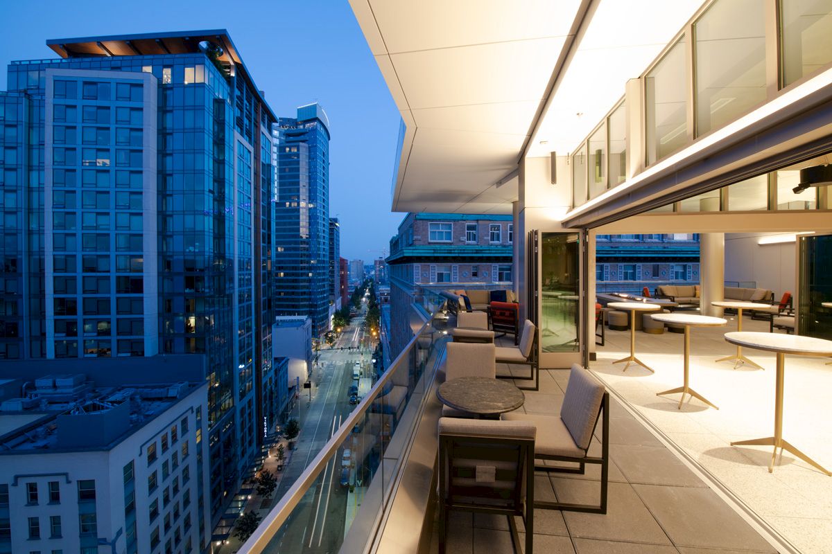 A modern urban balcony scene at dusk, with seating, tables, and city buildings in the background, under a blue evening sky.