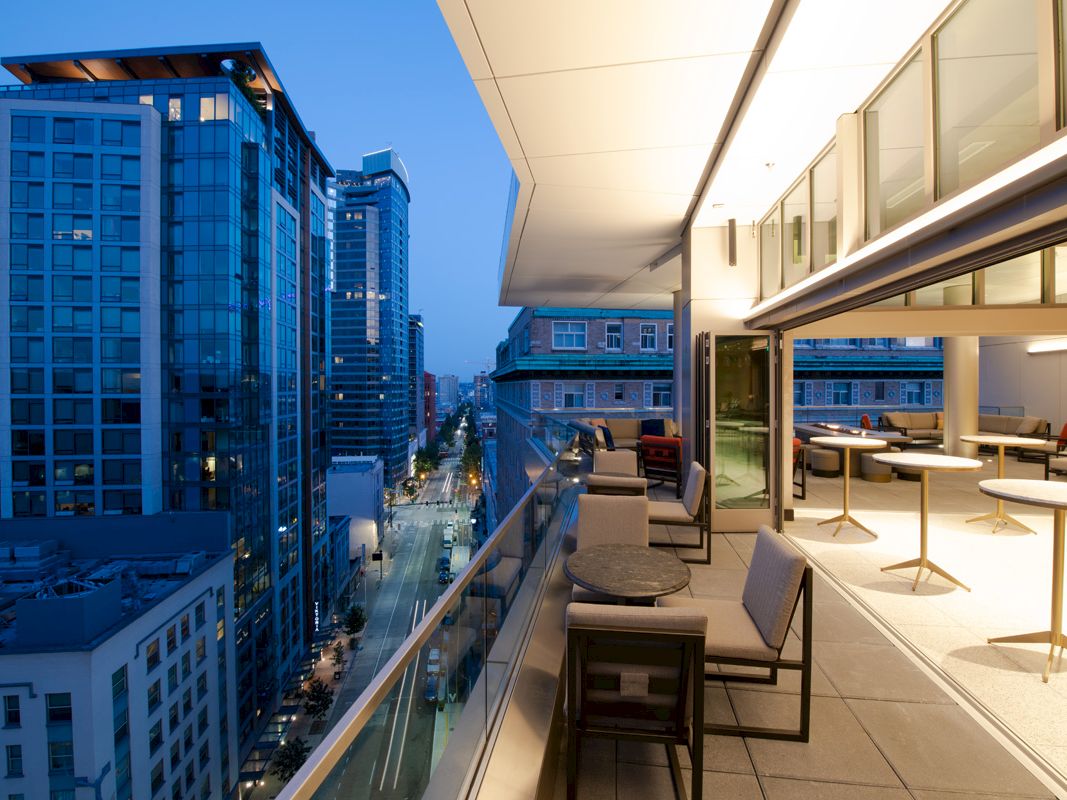 A modern urban balcony scene at dusk, with seating, tables, and city buildings in the background, under a blue evening sky.