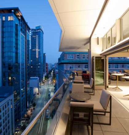 Rooftop balcony with tables and chairs overlooks a city street lined with modern buildings during twilight.