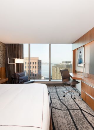 A modern hotel room with a bed, desk, chair, and large TV, featuring a window view of city buildings and water in the background.