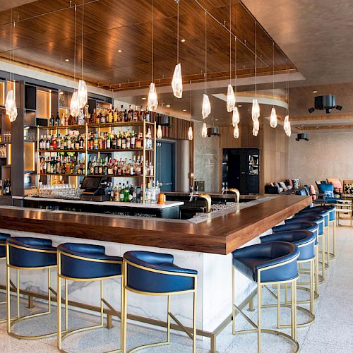 The image shows a modern bar with a wooden counter, blue bar stools, hanging lights, and a well-stocked liquor display.