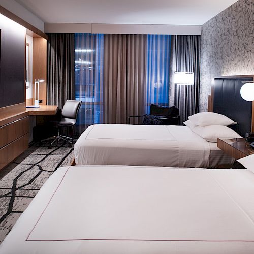 The image shows a modern hotel room with two beds, a TV, a desk, and stylish decor, featuring a patterned wall and curtains.