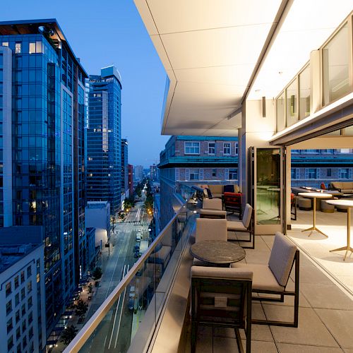 An outdoor terrace with seating overlooks a city street lined with tall modern buildings during evening time.