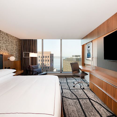 A modern hotel room with a large bed, wall-mounted TV, desk, and a city view through large windows.