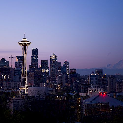 A cityscape at dusk with a distinctive tower, skyscrapers, and a mountain in the background under a clear sky.