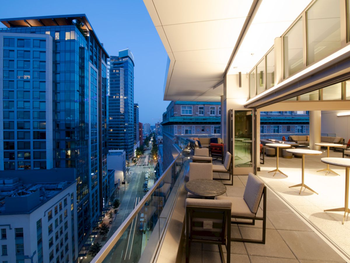 The image shows a modern urban balcony overlooking a city street with tall buildings, outdoor seating, and warm lighting at twilight.