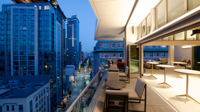 The image shows a modern urban balcony overlooking a city street with tall buildings, outdoor seating, and warm lighting at twilight.