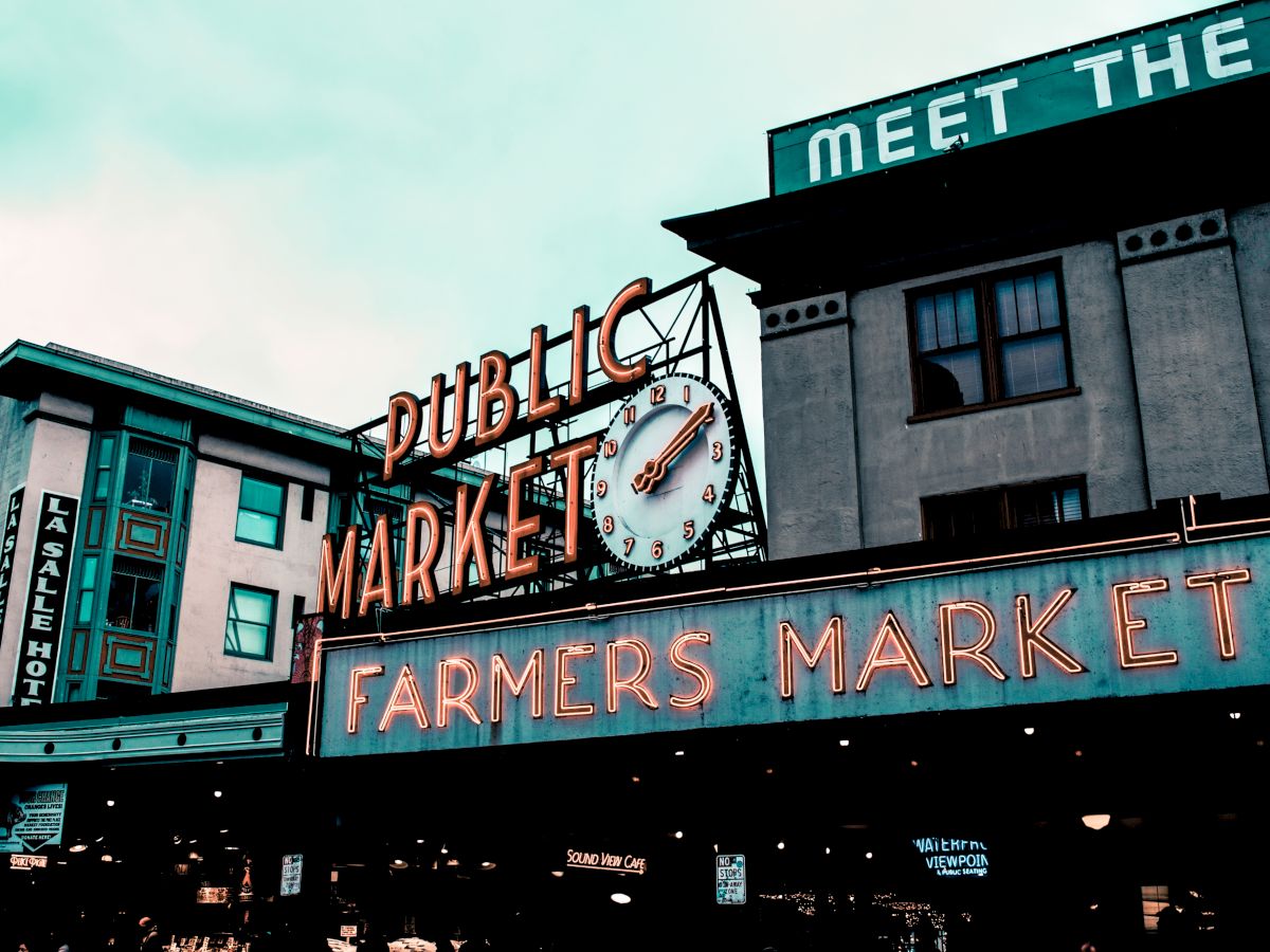 The image shows the iconic neon signs for a public market and farmers market, with a clock, set against a building backdrop.