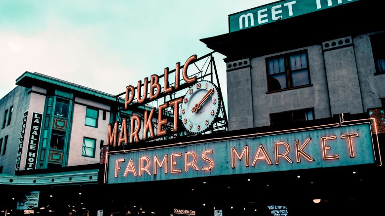The image shows the iconic neon signs for a public market and farmers market, with a clock, set against a building backdrop.