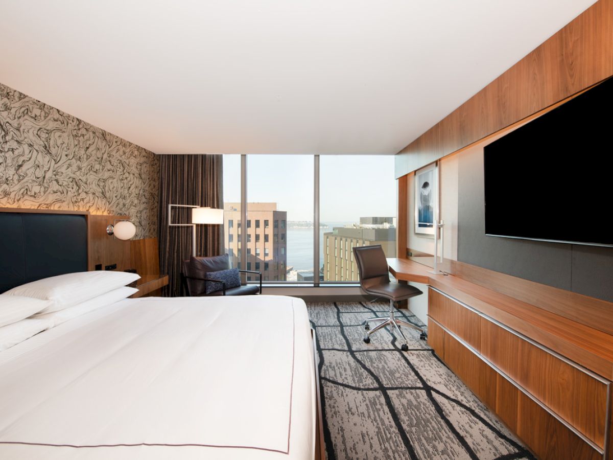 A modern hotel room features a large bed, wall-mounted TV, desk with chair, and a window with a city view.