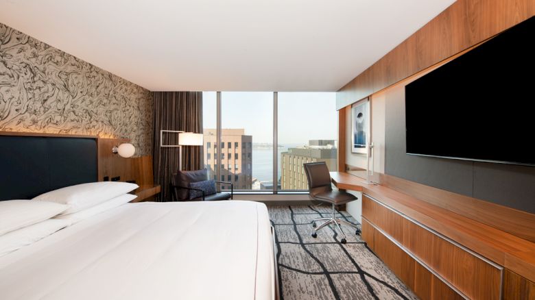 A modern hotel room features a large bed, wall-mounted TV, desk with chair, and a window with a city view.
