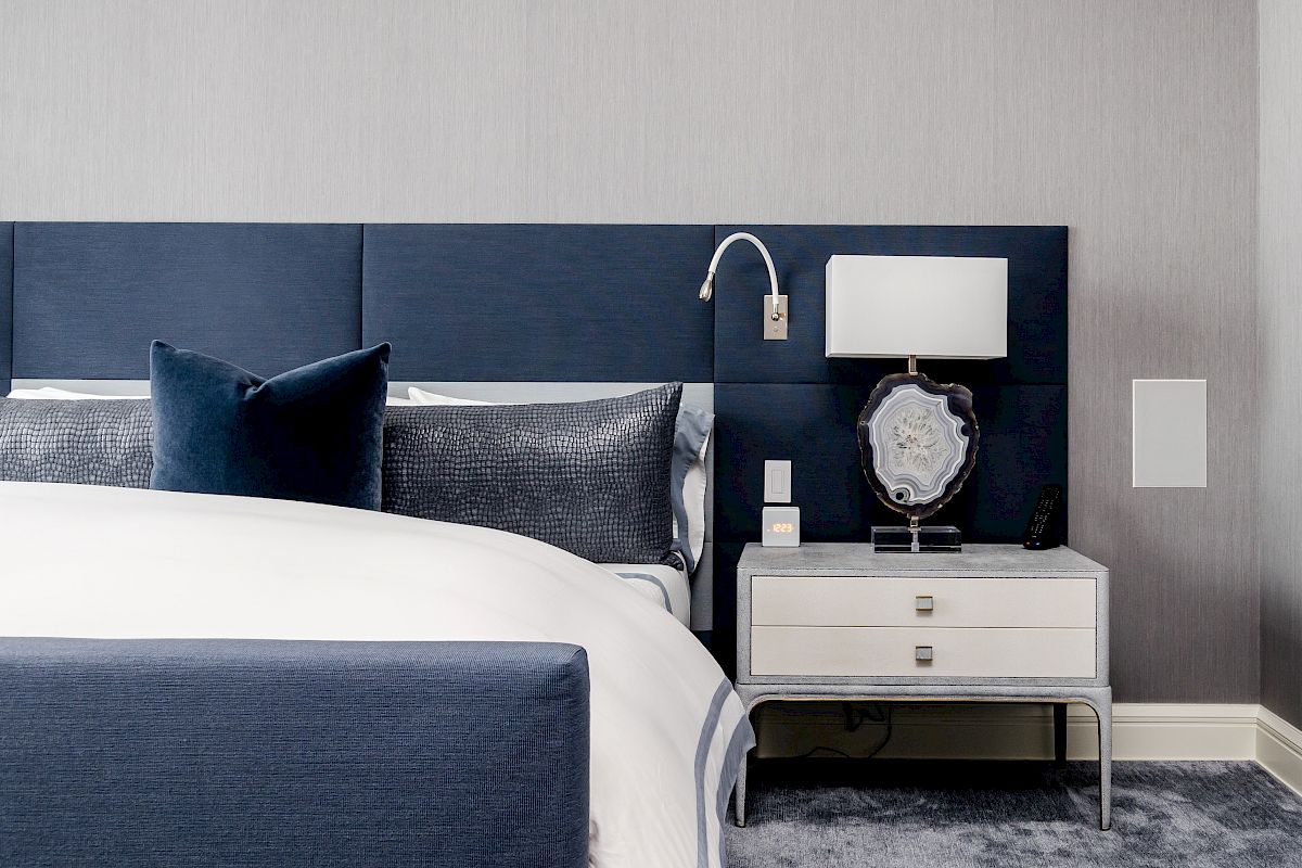 A modern bedroom with a blue headboard, white bedding, a nightstand, and a lamp, in a minimalist design.