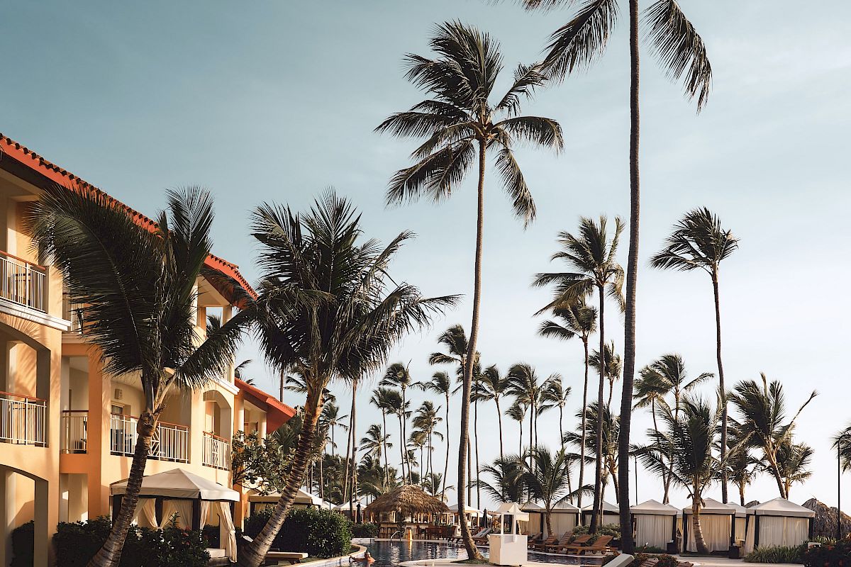 A resort poolside with palm trees, sun loungers, and an adjacent building, under a clear blue sky.