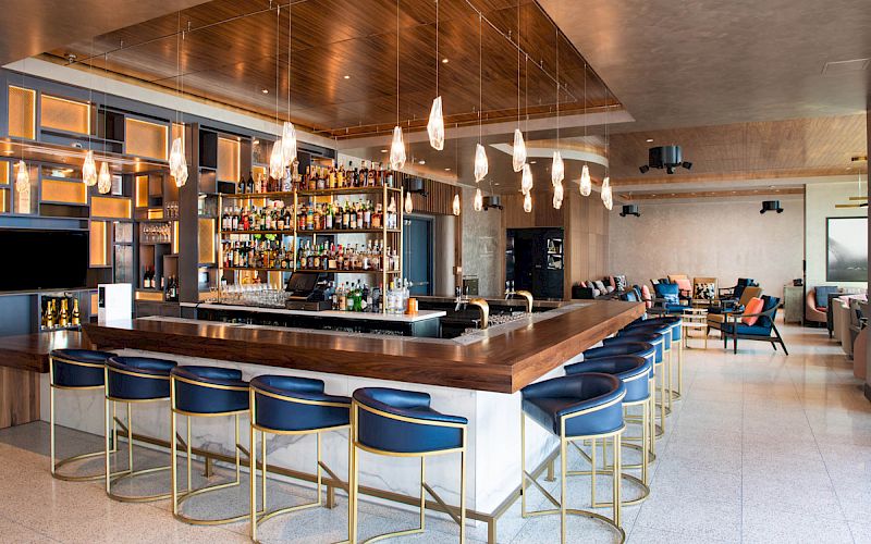 A modern bar with blue stools, a well-stocked liquor shelf, pendant lights, and seating area in a stylish, open space.