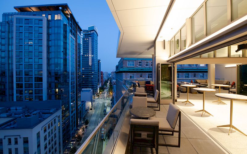 A modern rooftop terrace with seating overlooks a city street lined with tall buildings at twilight.