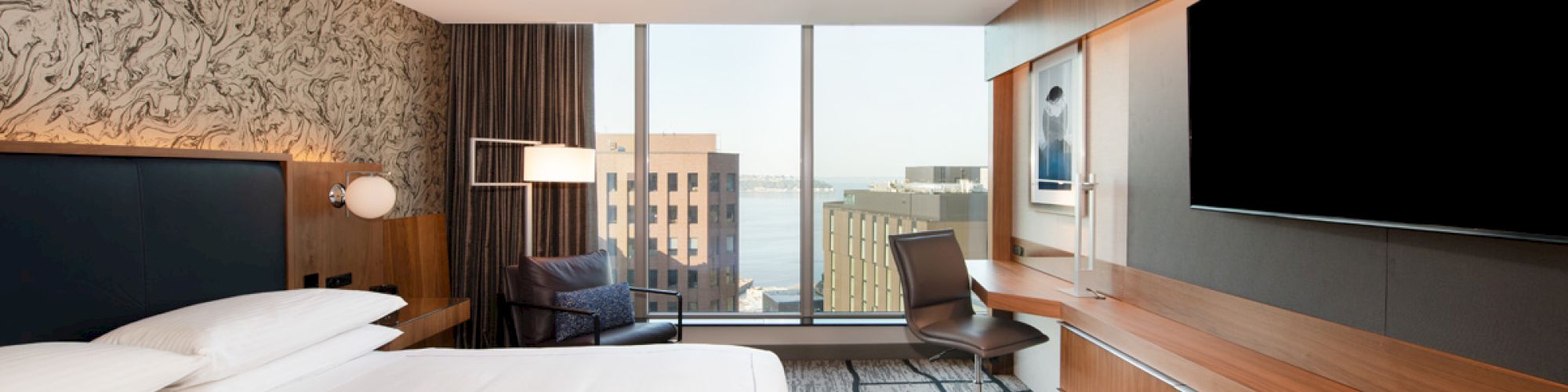 A modern hotel room with a large bed, wall-mounted TV, desk, chair, and a window offering a cityscape view.